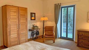 Ein Schlafzimmer mit einem großen Bett, einem Holzschrank, einem Sessel, einem kleinen Tisch mit Lampe und Blick durch eine Glastür.