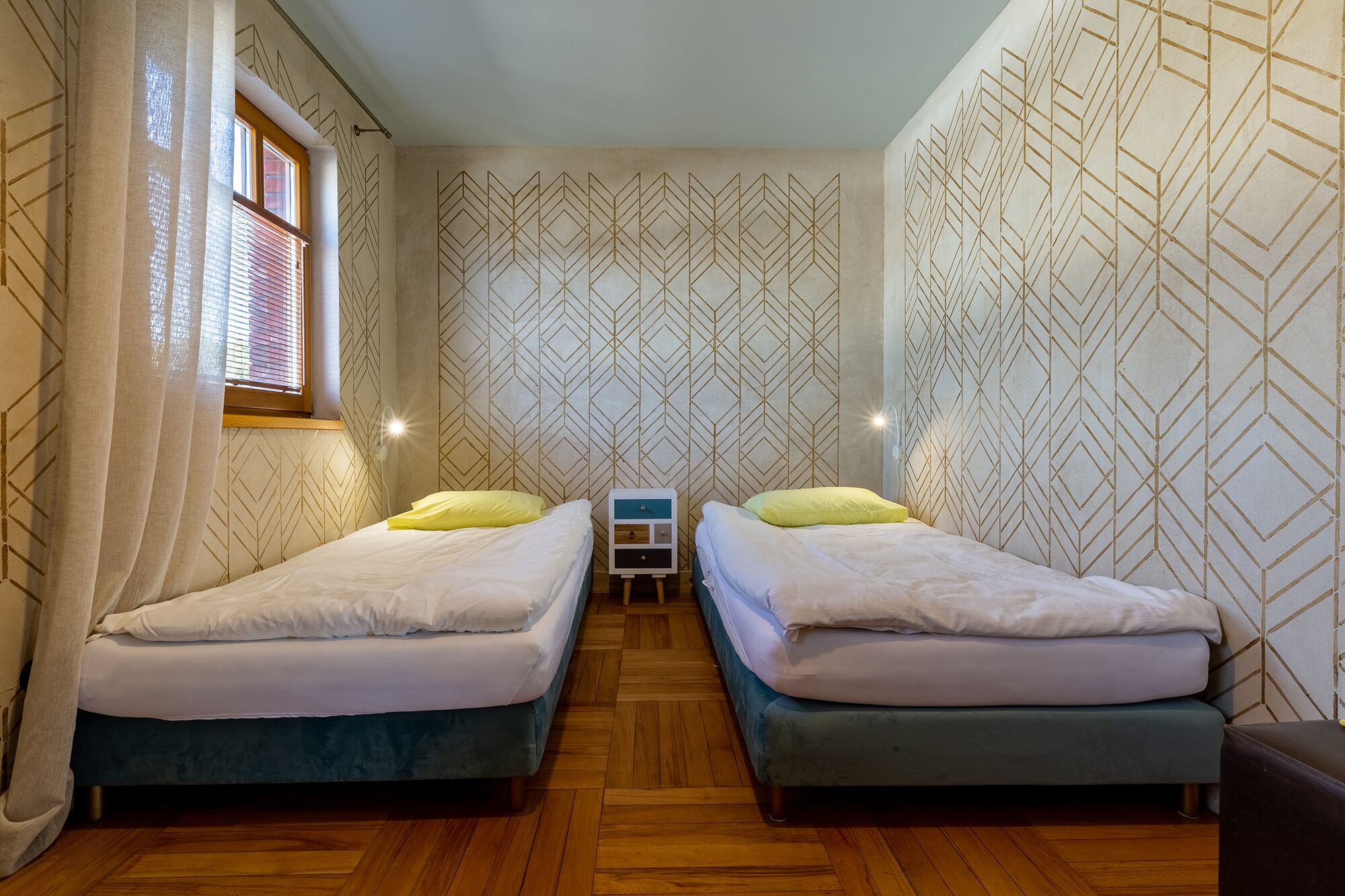 A room with two beds, a window with blinds, patterned wallpaper, and a small bedside table with a lamp.