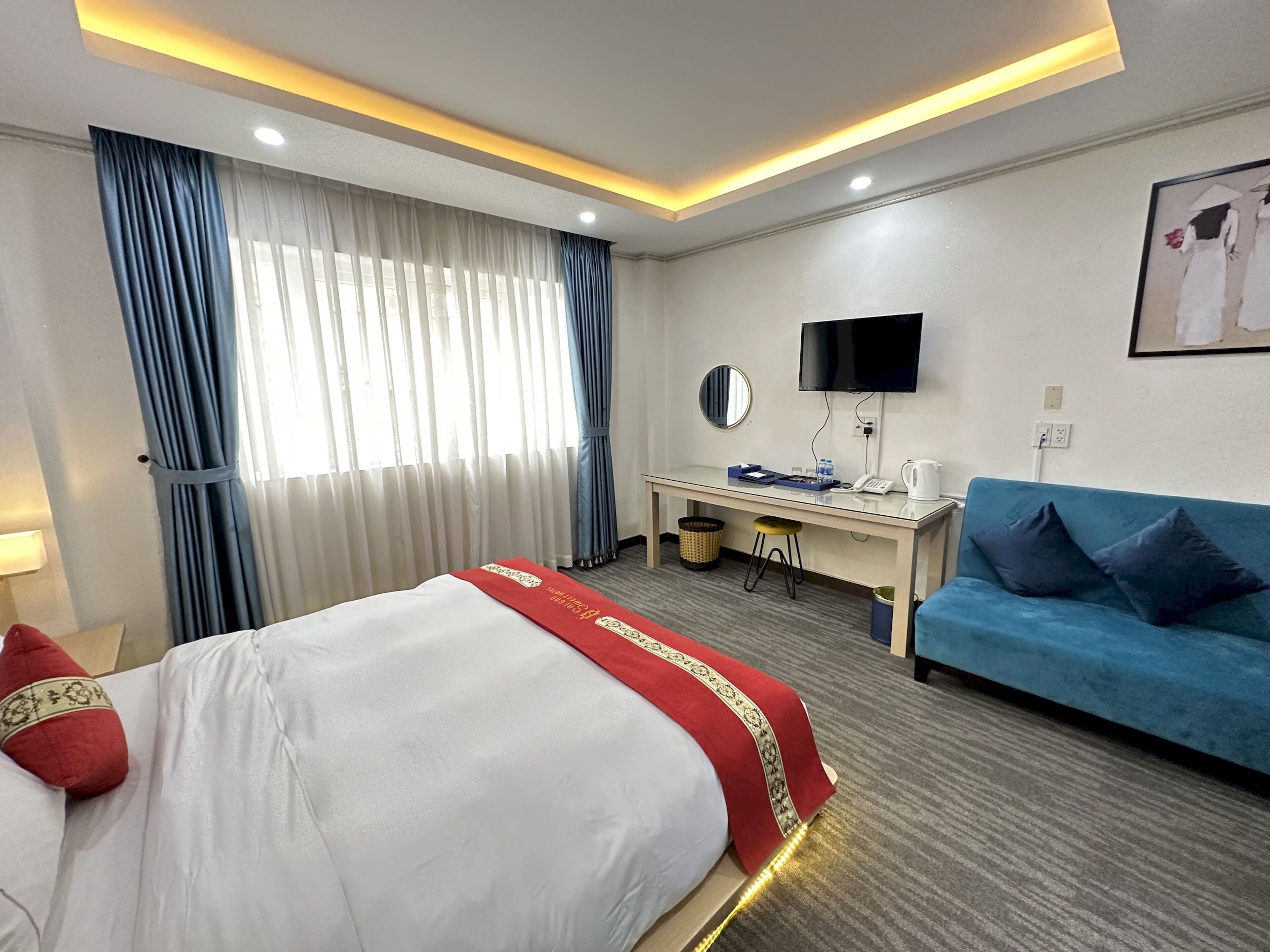 Una habitación de hotel con cama, escritorio, sofá azul, televisor y ventana con cortinas.