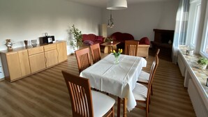 Ein Essbereich mit einem Holztisch, Stühlen und einer weißen Tischdecke. Im Hintergrund steht ein Holzschrank, ein Sofa und eine Pflanze.
