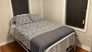 A single bed with a patterned bedspread and pillows, two windows with blinds, and a white metal bed frame. - Maggie's Place (Lookout Mountain)
