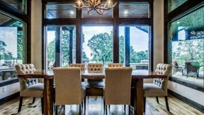 Zona pranzo con tavolo in legno e sei sedie imbottite, ampie finestre vista alberi e un lampadario.