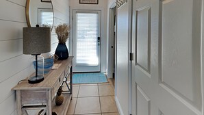 A hallway with a wooden bench, a lamp, a vase with dried plants, a blue mat, and a door with a sign above it. - Bonita Point Condo II #214E by Resort Collection (Panama City Beach)