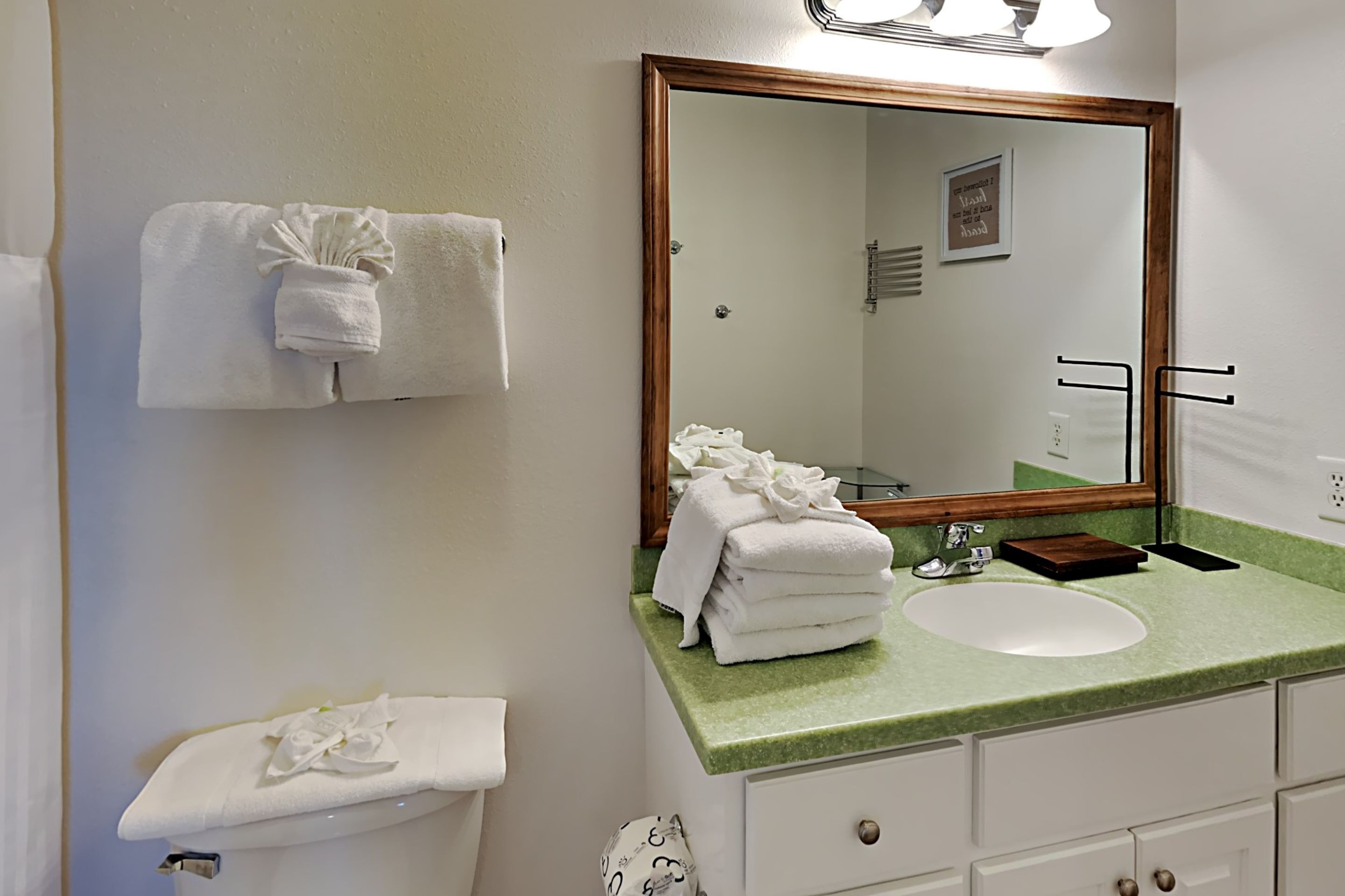 A bathroom with a green countertop, white sink, and a large mirror. There are towels hanging on the wall and a small wooden tray on the countertop.