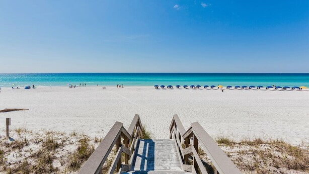 A beach with a wooden staircase leading down to the sand, a clear blue sky, and a row of beach umbrellas. - Bonita Point Condo II #214E by Resort Collection (Panama City Beach)