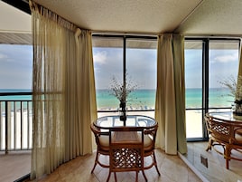 A balcony with a view of the beach, a round table with chairs, and a vase with flowers.
