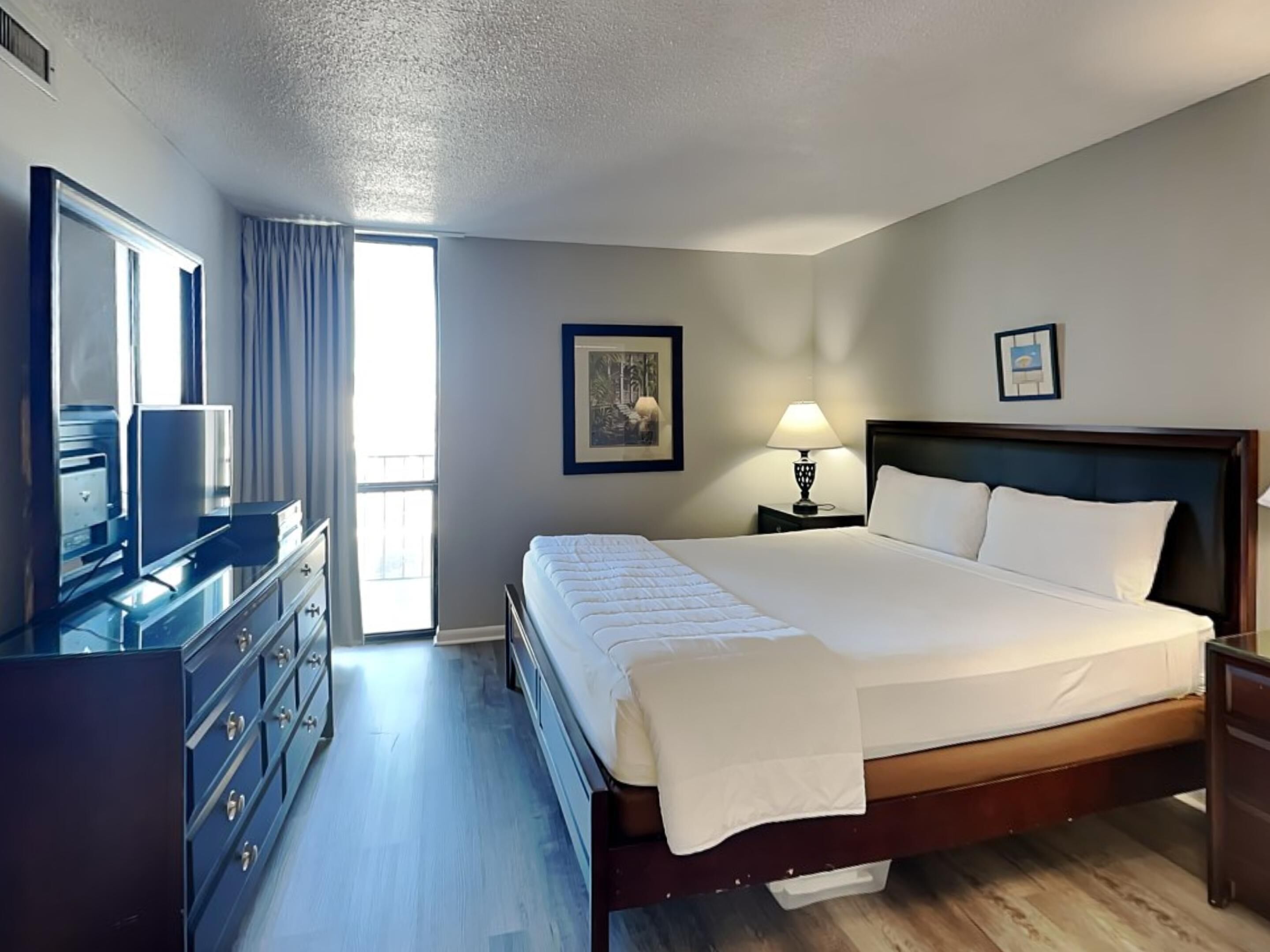 A hotel room with a large bed, a dresser, a television, and a window with curtains.