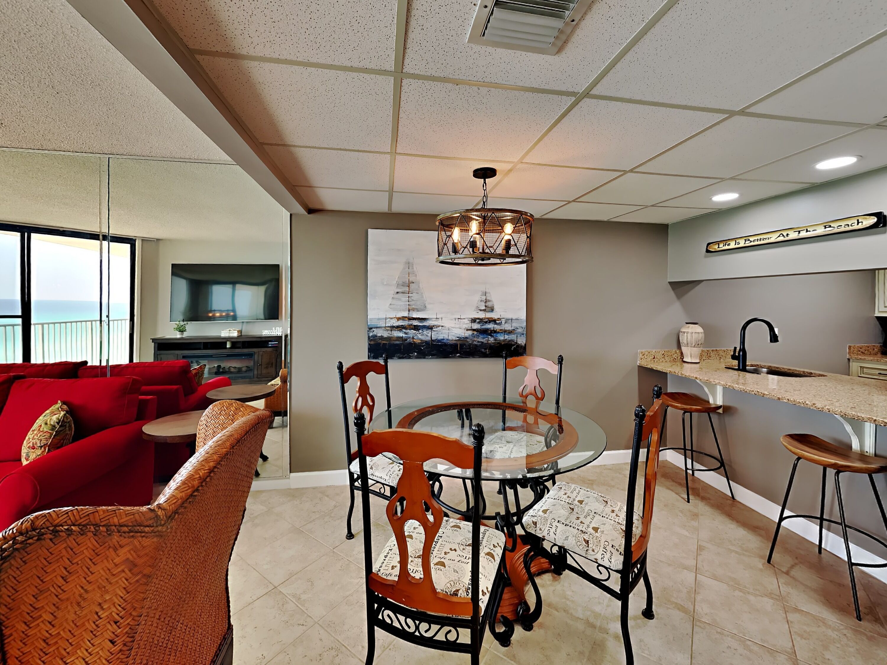 A modern living room with a red sofa, wicker chairs, a glass dining table, and a kitchen area with a countertop and stools.