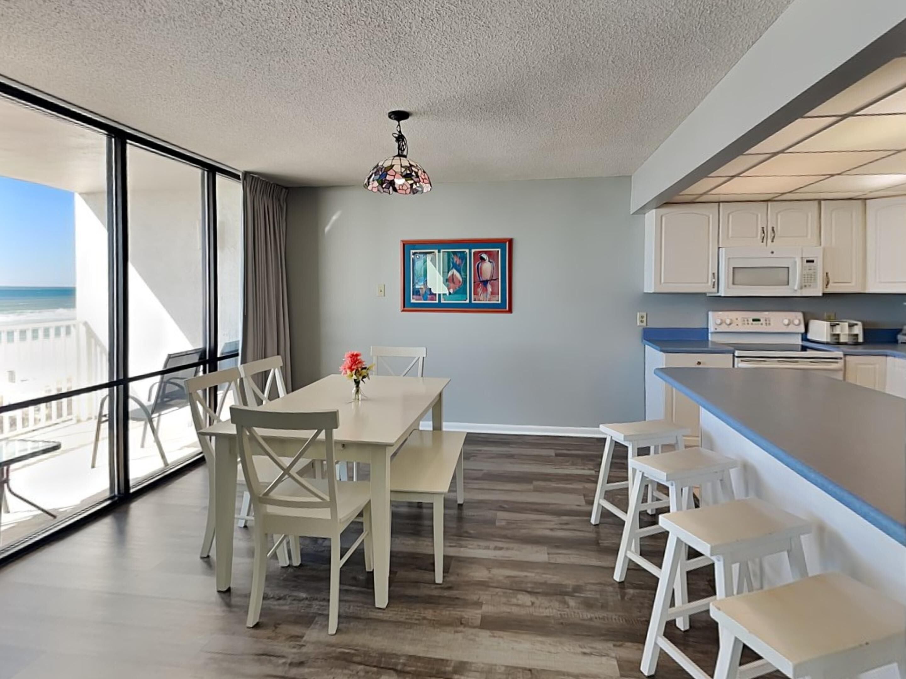 A modern kitchen with white cabinets and a blue countertop, a dining area with white chairs and a table, and a large window overlooking the ocean.