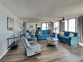 A spacious living room with a blue sofa, a leather armchair, and a coffee table. There is a dining area in the background with a table and chairs.