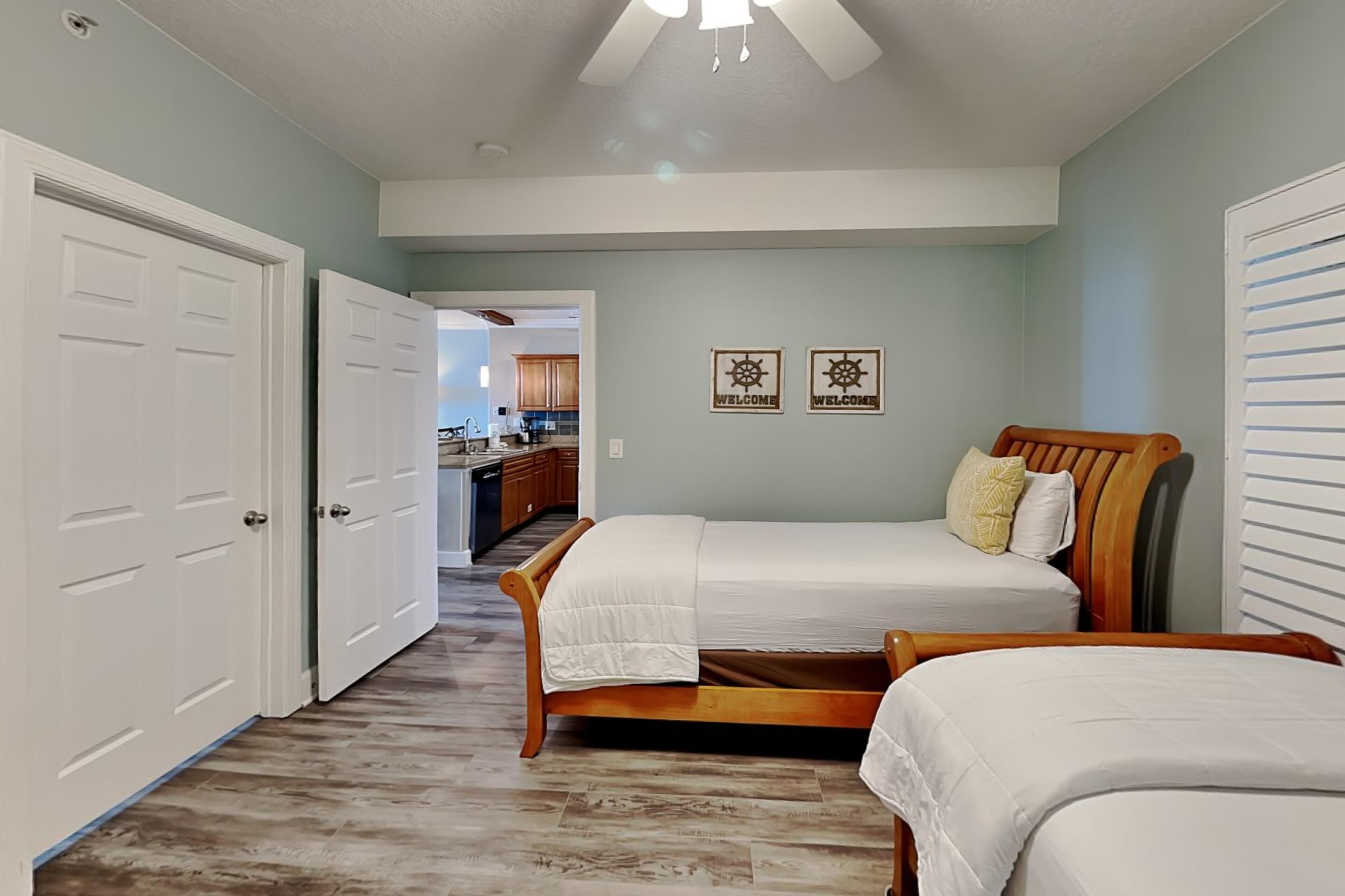 A bedroom with two beds, a wooden headboard, a ceiling fan, and a kitchen in the background.