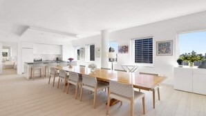 Un vaste espace dédié à la salle à manger, avec une longue table en bois et des chaises, une cuisine en arrière-plan et de grandes fenêtres offrant une vue dégagée.