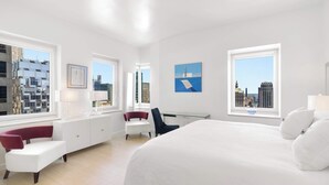 Une chambre d’hôtel moderne, dotée d’un grand lit, d’un bureau et d’une chaise. Deux fenêtres offrent une vue sur la ville.