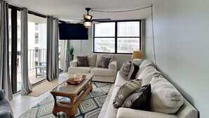 Un salon avec un canapé, une table basse et un téléviseur. Il y a un balcon avec des rideaux et une plante.