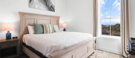 A bedroom with a large bed, a headboard, two bedside tables with lamps, a window with curtains, and a painting on the wall.