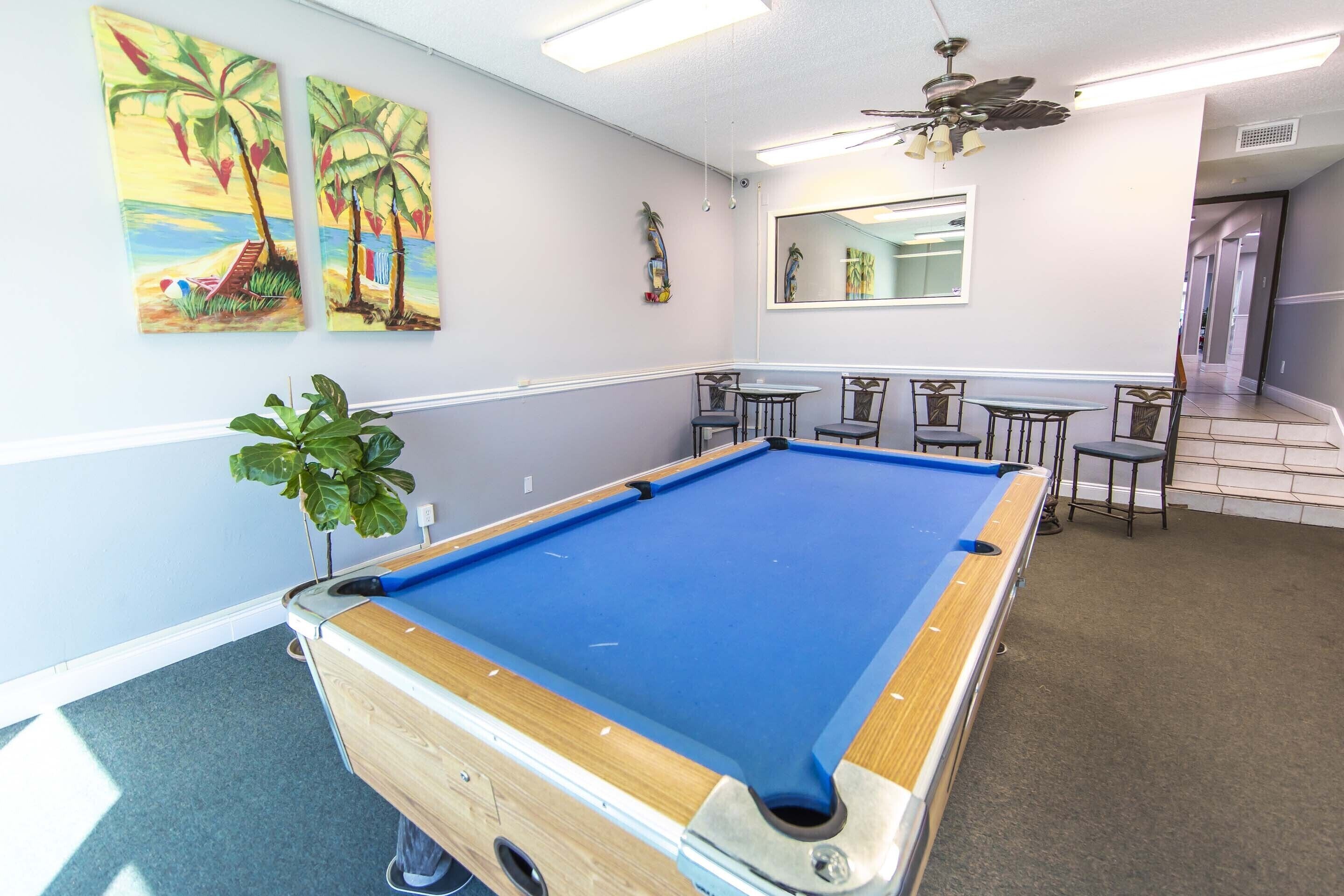 A pool table in a room with a ceiling fan, a large mirror, and two paintings of palm trees.