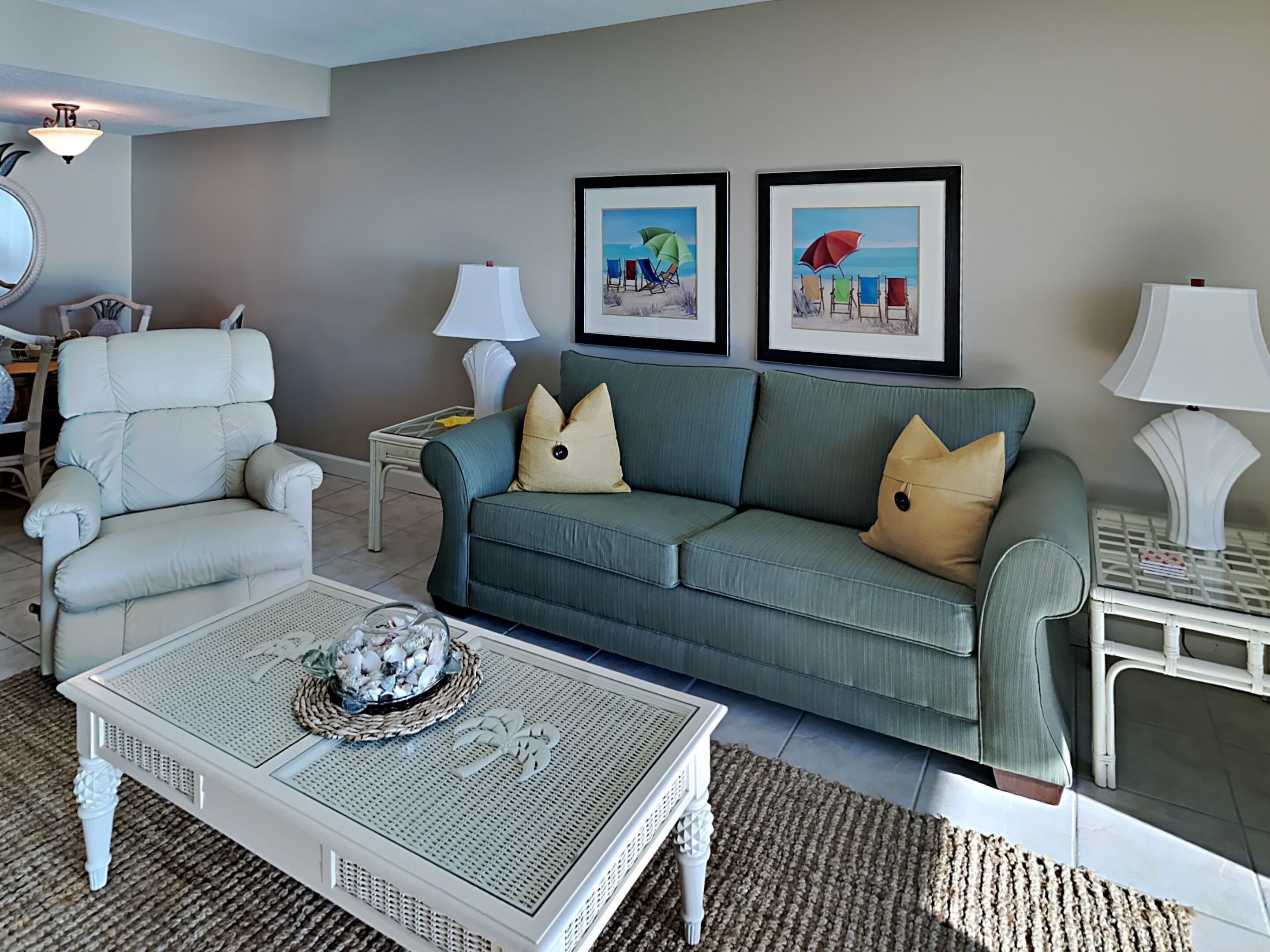 A living room with a grey sofa, a white armchair, and a coffee table with decorative items. There are two framed pictures on the wall, a lamp on a side table, and a rug on the floor.
