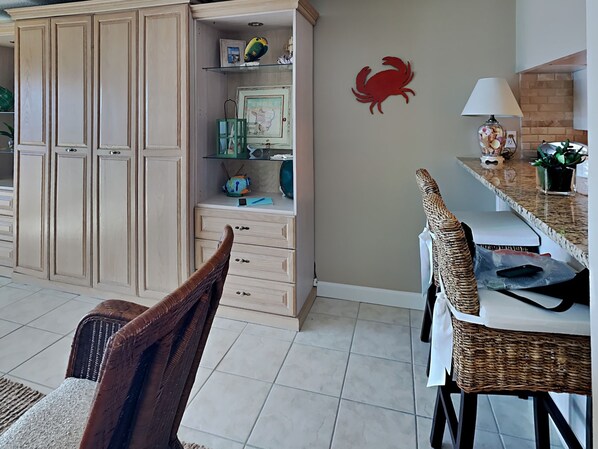 A kitchen with a dining area, featuring a cabinet, a wicker chair, a table, and a red crab wall decoration.