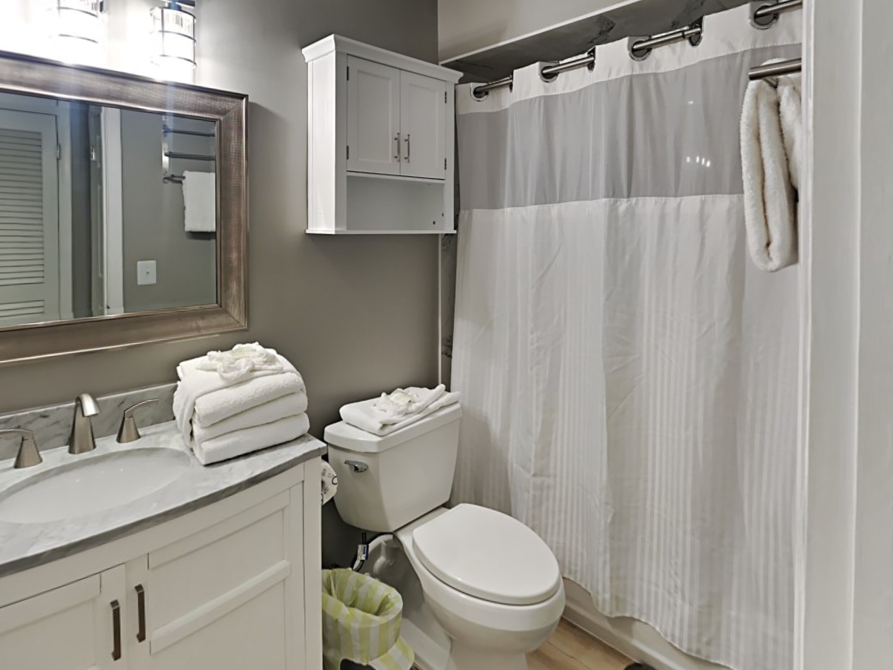 Une salle de bain avec un lavabo blanc, des toilettes et un rideau de douche. Des serviettes sont pliées sur le lavabo et une poubelle se trouve sous les toilettes.