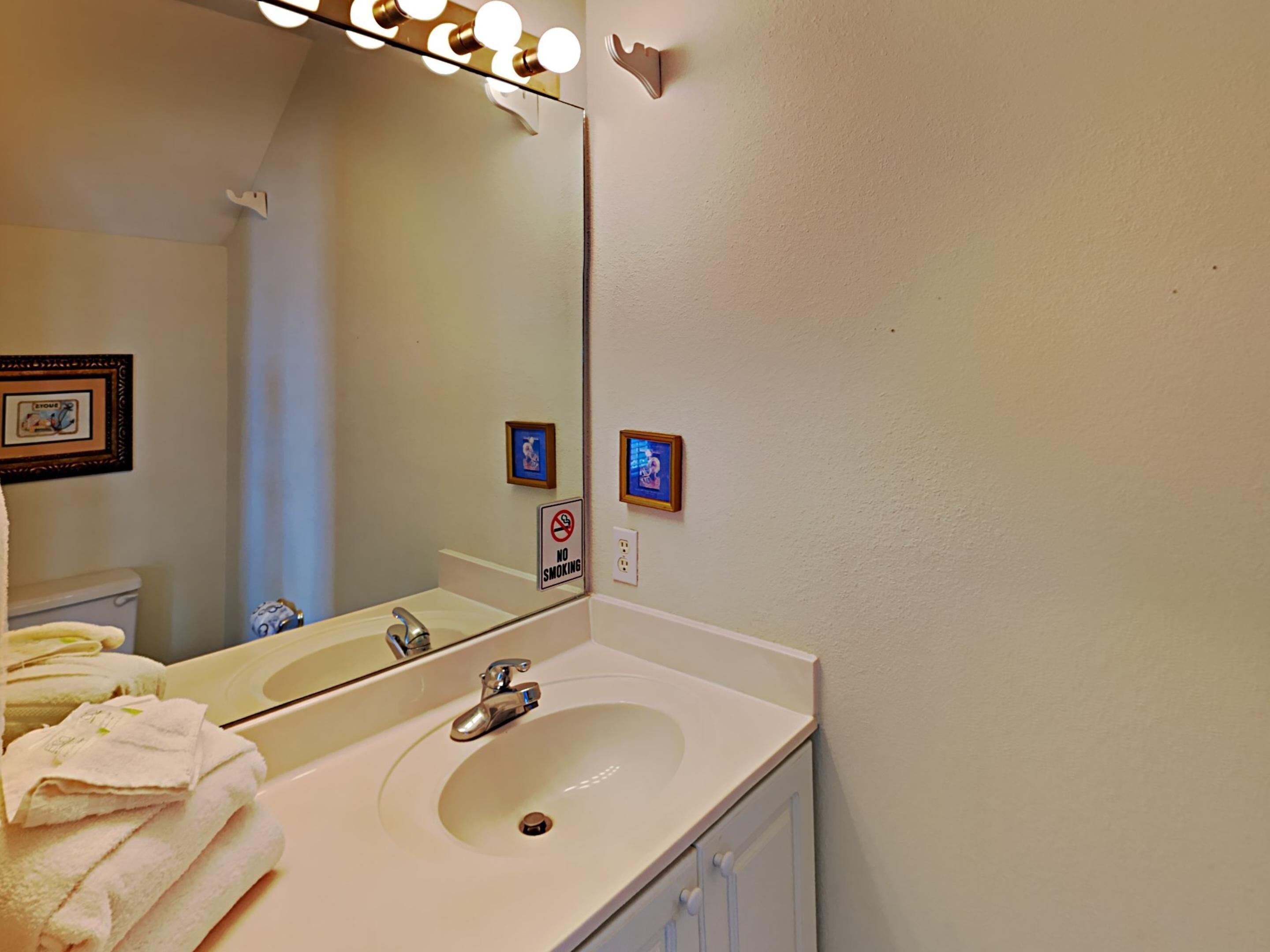 A bathroom with a sink, mirror, and lights above the mirror. There are towels stacked near the sink and a 'No Smoking' sign on the wall.