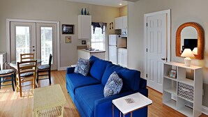 A living room with a blue sofa, a small white table, and a wicker chair. There is a kitchen area with cabinets and a refrigerator in the background. - Seahawk South B by Southern Vacation Rentals (Gulf Shores)