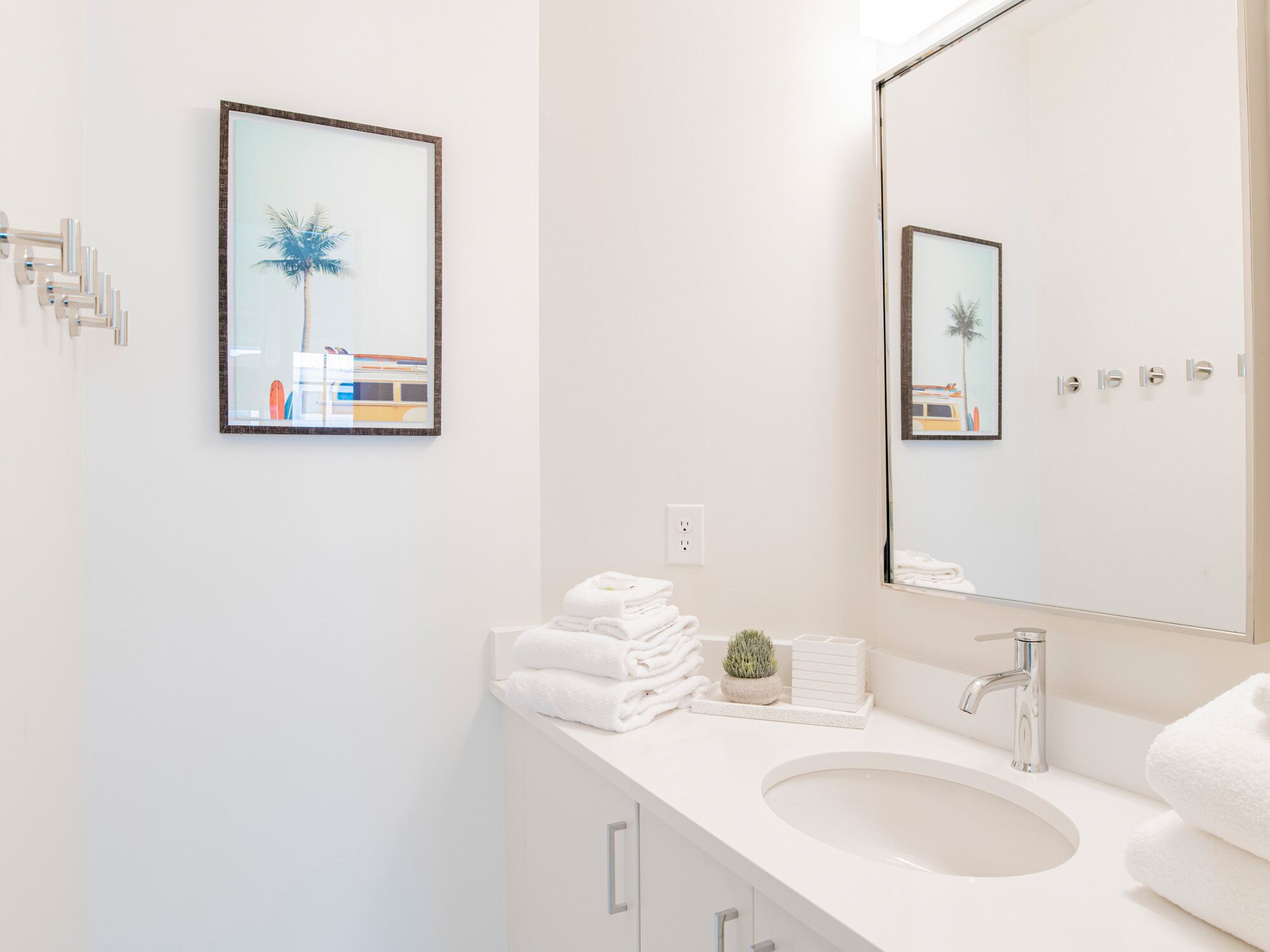 A modern bathroom with a white sink, a large mirror, and a framed picture of a palm tree and train on the wall.