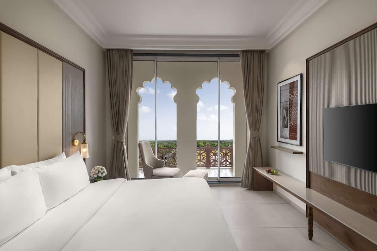 A hotel room with a large bed, a TV, a chair, a balcony with a view, and a desk with a vase.