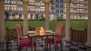 Un comedor con una mesa dispuesta para dos personas, rodeado de sillas ornamentales, y vista a un edificio grande de varios pisos al fondo.