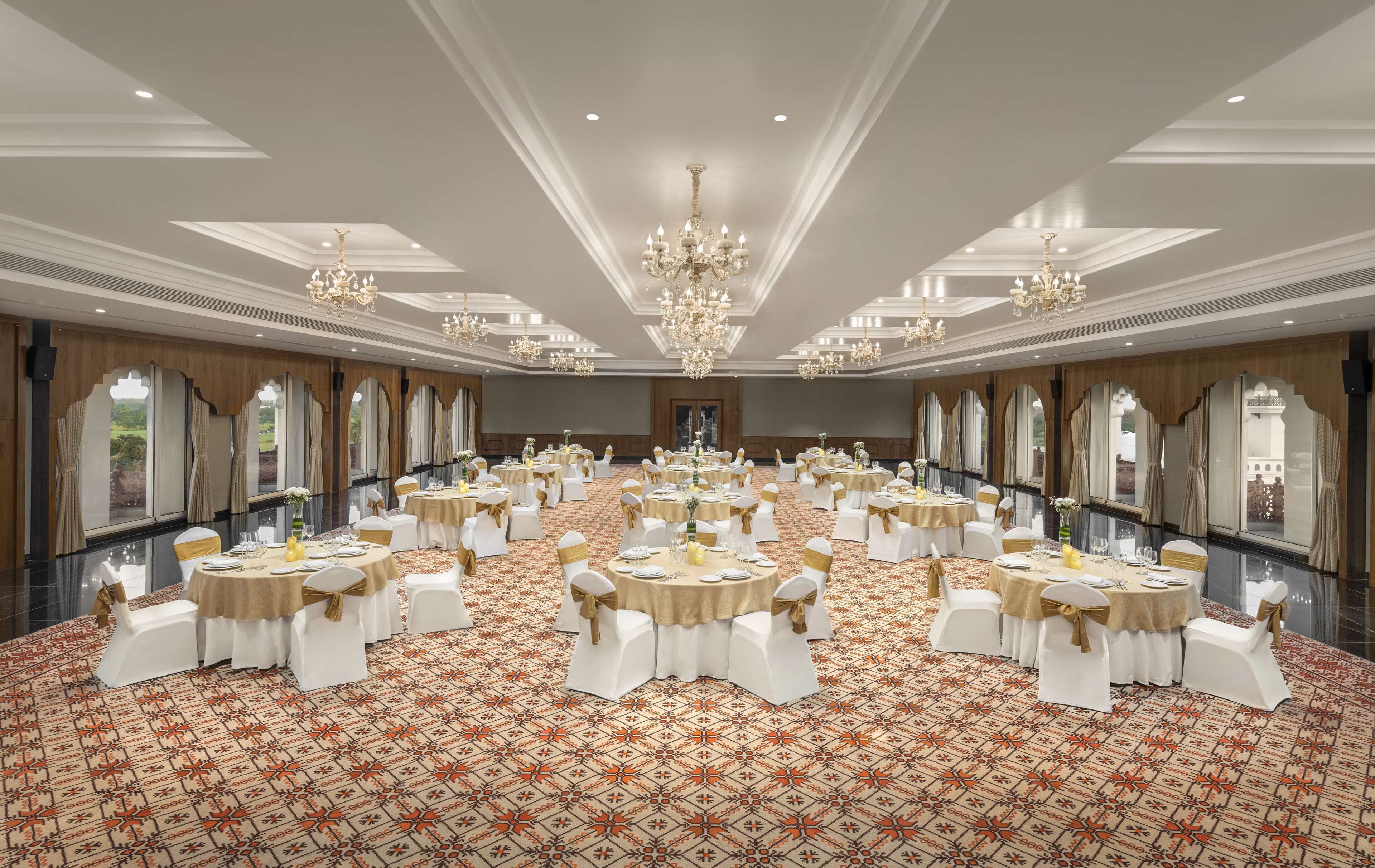 A large banquet hall with round tables, white chairs, chandeliers, and patterned carpet.