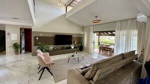 Un'ampia zona giorno con un divano grande, una poltrona rosa, un tavolino da caffè e una TV a parete. Include un balcone con tavolo e sedie, e vista sul verde.