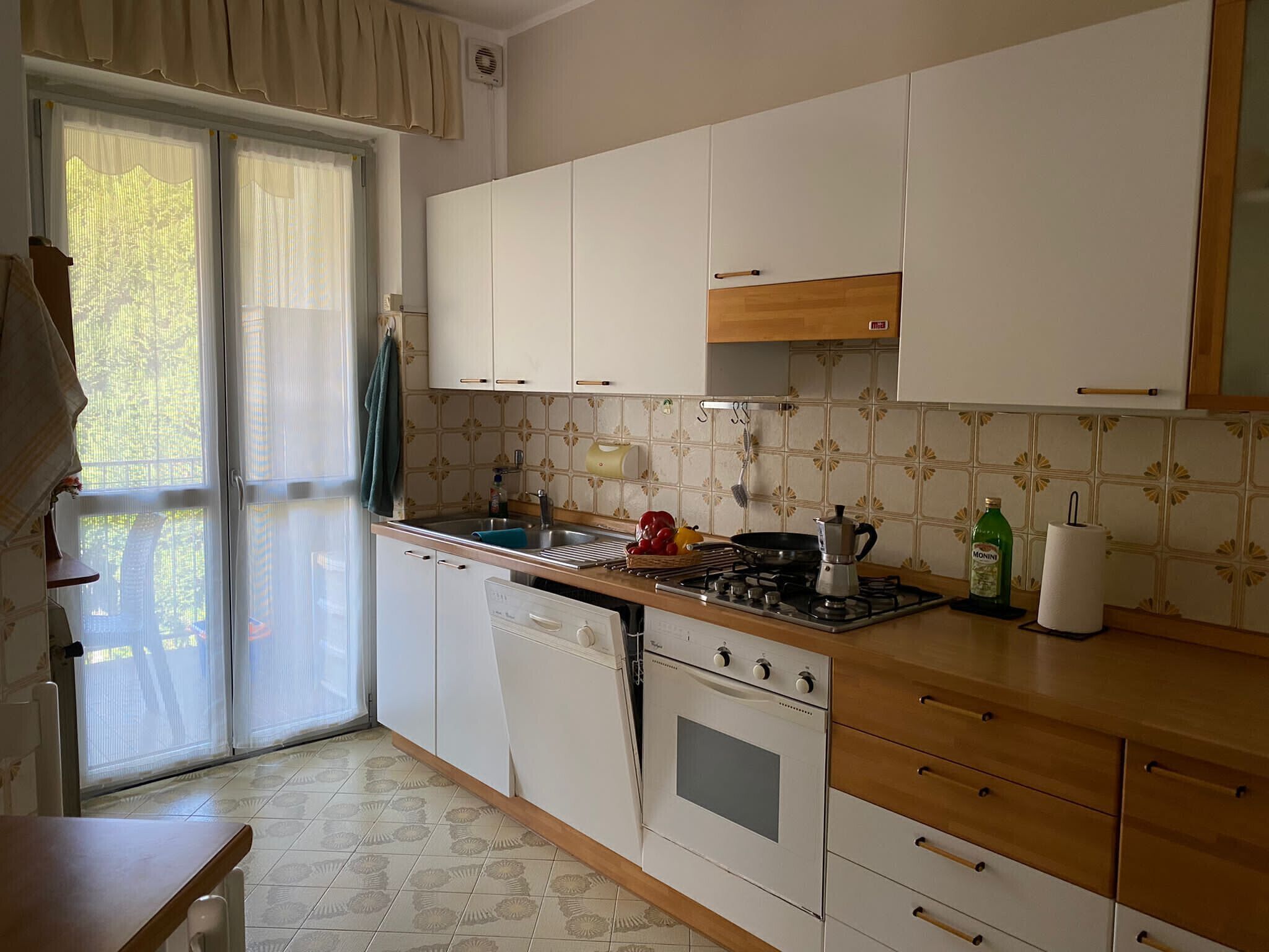 Cucina con mobili bianchi, fornello, lavello e finestra con tende.