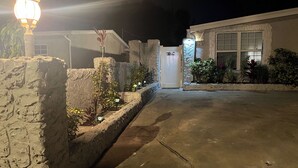 Vue nocturne d’une allée menant à une résidence, avec un mur en pierre, un lampadaire allumé et une maison à la façade en pierre.