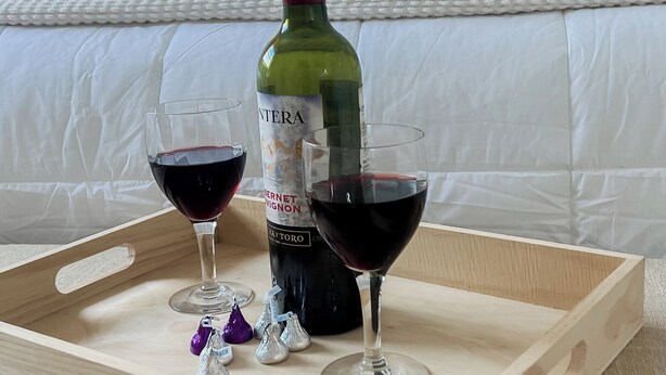 Un plateau en bois avec une bouteille de vin et deux verres de vin rouge, posé sur un lit recouvert de draps blancs, avec une peinture florale en arrière-plan.