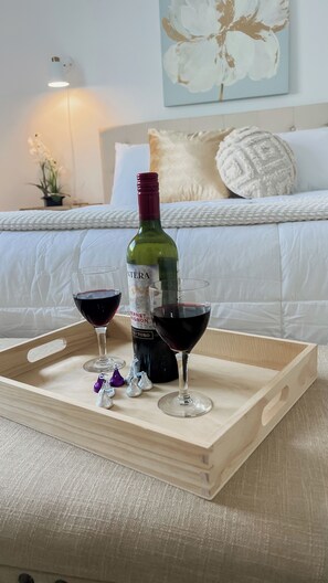 Una bandeja de madera con una botella de vino y dos copas de vino tinto, sobre una cama con ropa de cama blanca y un cuadro floral de fondo.