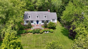 A two-story house with a front porch, surrounded by lush greenery and trees.