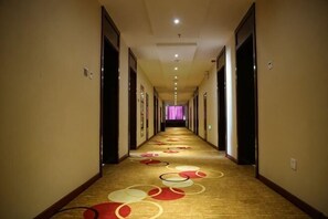 Um corredor de hotel com piso revestido de carpete, portas em ambos os lados e um longo vão que leva a um quarto com janela iluminada em roxo.
