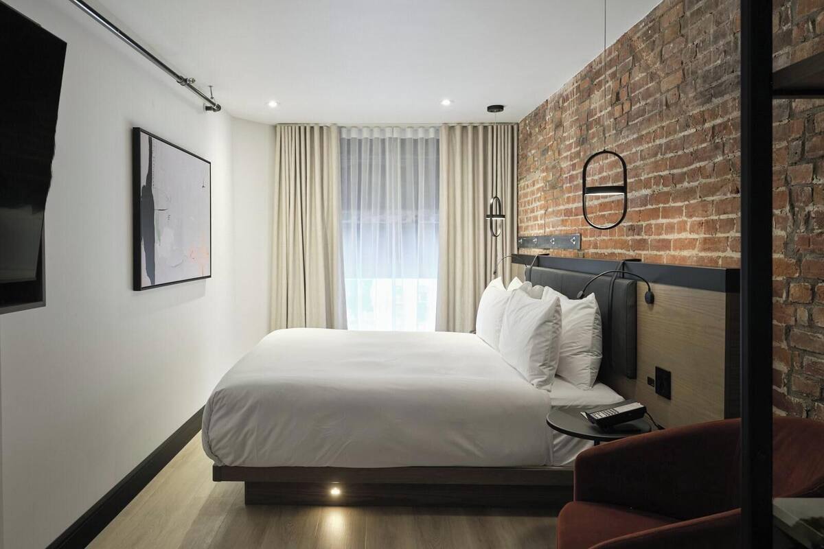 Une chambre d’hôtel moderne dotée d’un grand lit, d’un mur décoratif en briques, d’un téléviseur à écran plat et d’une fenêtre avec des rideaux.