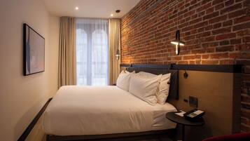 A hotel room with a large bed, a brick wall, a window with curtains, a small table with a phone, and a red chair.