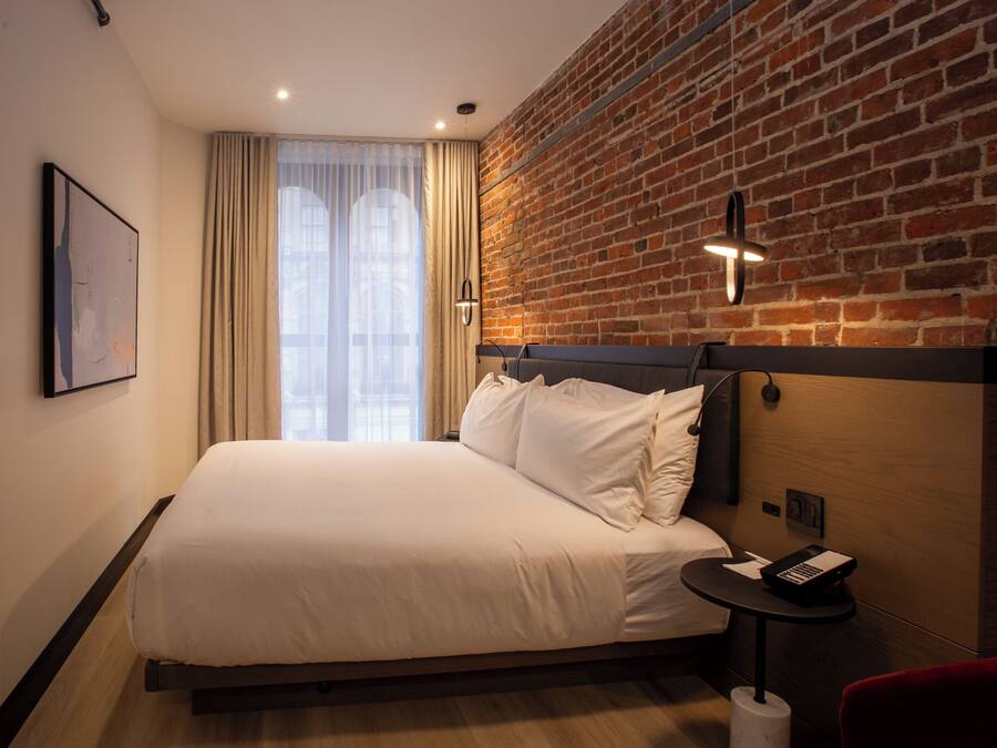A hotel room with a large bed, a brick wall, a window with curtains, a small table with a phone, and a red chair.
