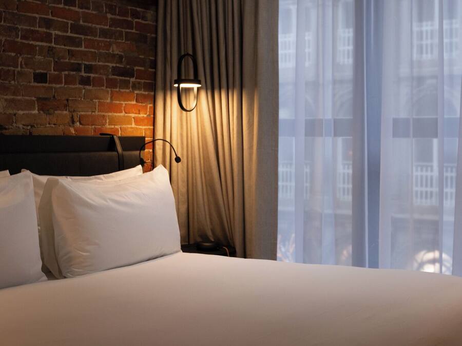 A hotel room with a brick wall, a bed with white linens, a wall-mounted lamp, and a window with sheer curtains.