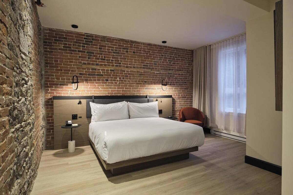 Une chambre d’hôtel avec un mur en briques, un grand lit avec du linge de lit blanc, une table de chevet avec une lampe et une chaise.