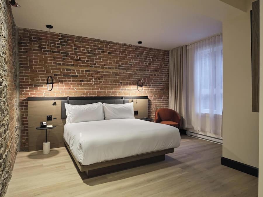 Habitación de hotel con pared de ladrillo, una cama grande con ropa blanca, una mesita de noche con lámpara y una silla.