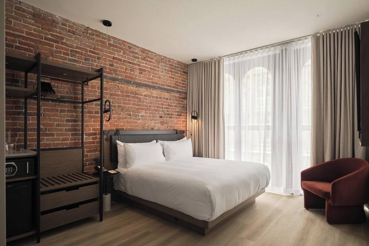 Une chambre d’hôtel moderne avec un mur en briques, un lit recouvert de draps blancs, une chaise rouge et un plancher en bois.