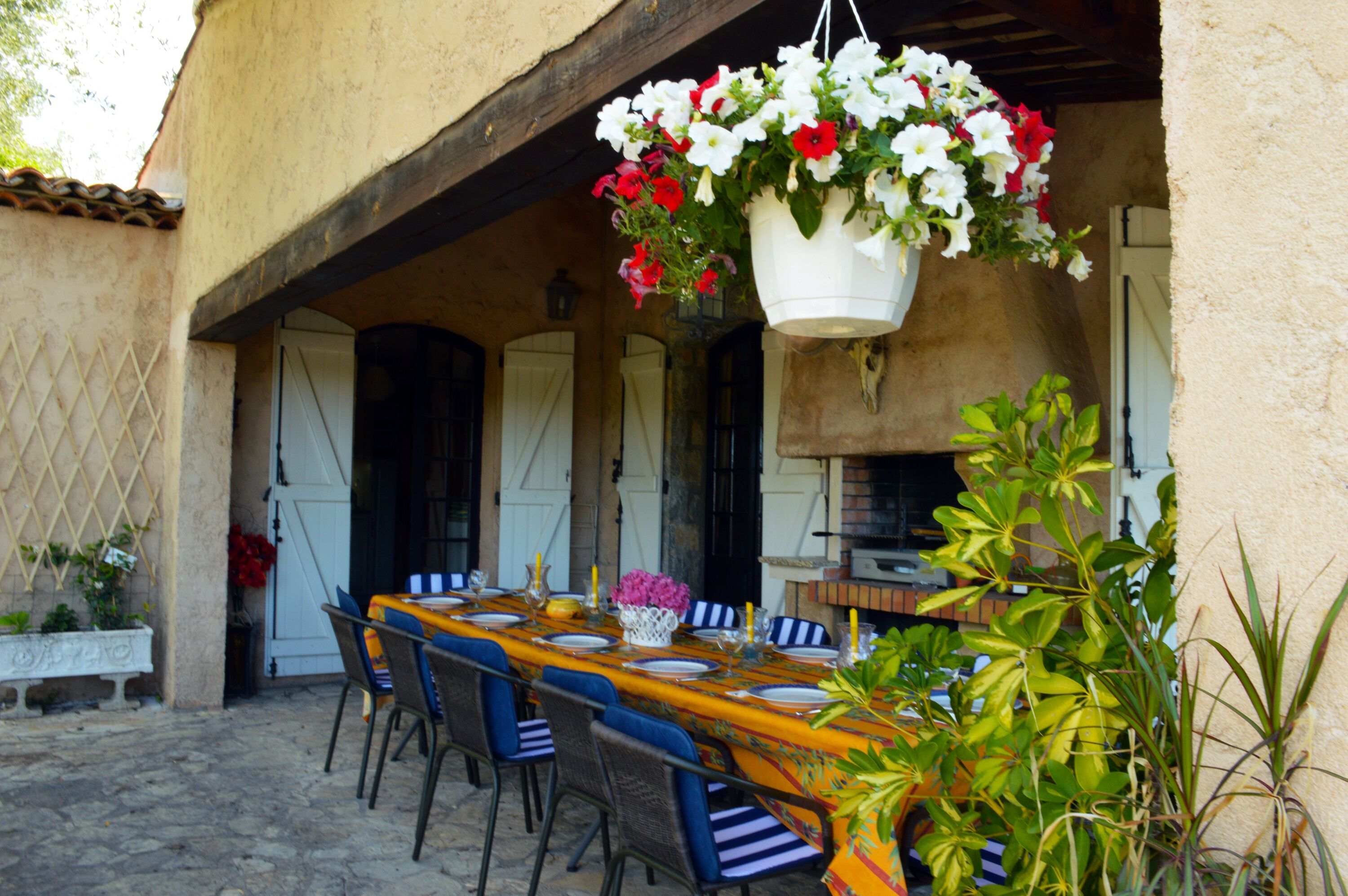 Et udendørs spiseområde med et bord dækket til fire personer, omgivet af stole, og en blomsterkurv hængende over.