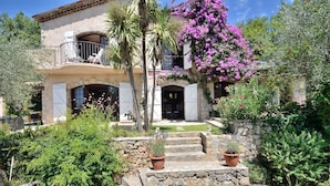 Eine mediterran gestaltete Villa mit Natursteinfassade, einem Balkon mit weißen Stühlen und einem üppigen Garten mit blühenden Pflanzen.