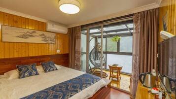 A bedroom with a large bed, a wooden headboard, a ceiling light, a window with curtains, and a small table with a chair.