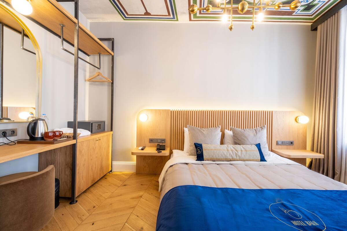 A modern hotel room with a wooden bed, bedside tables, a desk with a kettle, and a mirror.