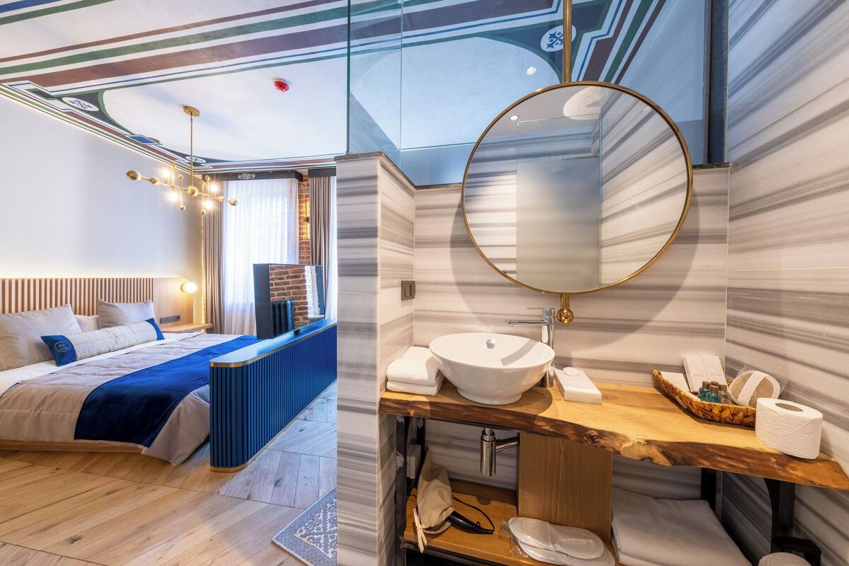 A modern hotel room with a large bed, a wooden headboard, a round mirror, a wooden vanity, and a blue accent wall.