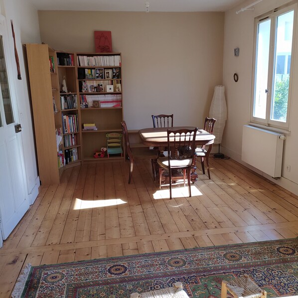 A room with a wooden floor, a dining table with chairs, a bookshelf, and a window.