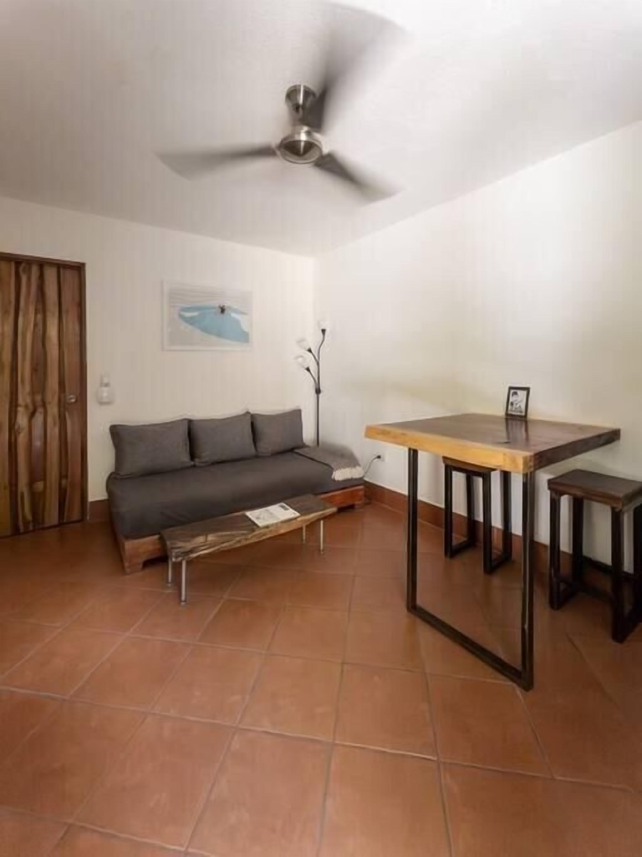 Ein Wohnzimmer mit grauem Sofa, einem Holztisch, einem kleinen Beistelltisch und einem Deckenventilator.